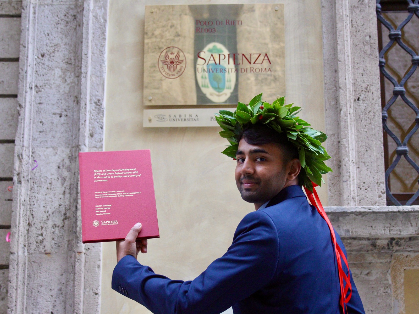 Usama with the laurel wreath, holding his bound thesis by the Sapienza plaque